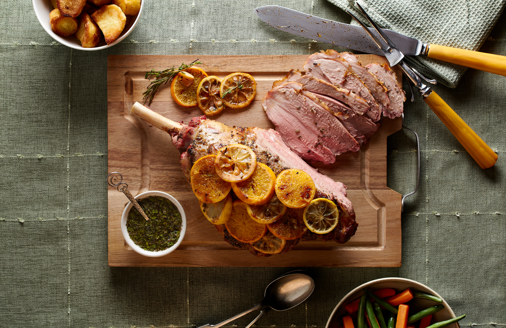 Large leg of lamb covered in orange slices on chopping board with carving knife and fork